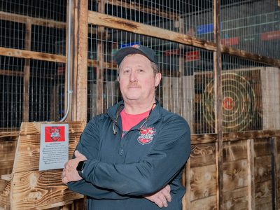 Man with arms folded in front of axe throwing bay.