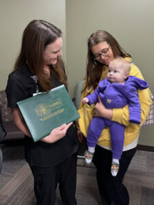 A smiling nurse in black scrubs holds a green folder labeled "The DAISY Award for Extraordinary Nurses" next to a young woman in a yellow hoodie holding a happy baby dressed in a purple outfit