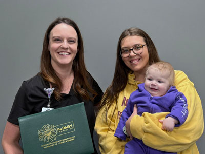 A smiling nurse in black scrubs holds a green folder labeled "The DAISY Award for Extraordinary Nurses" next to a young woman in a yellow hoodie holding a happy baby dressed in a purple outfit. The group is standing in front of a plain gray background.